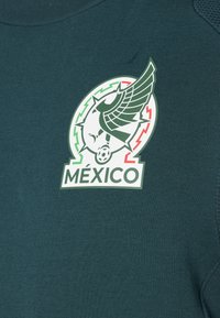 Πράσινο μπλουζάκι από ύφασμα με έντονο λογότυπο που απεικονίζει φτερωτό σχέδιο, μια ποδοσφαιρική μπάλα και τη λέξη "MÉXICO" σε λευκά κάτω από το έμβλημα.