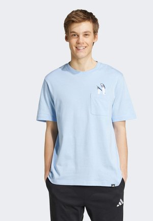 Young man smiling, wearing light blue t-shirt with small pocket and soccer cleats graphic above pocket, paired with black Adidas pants.