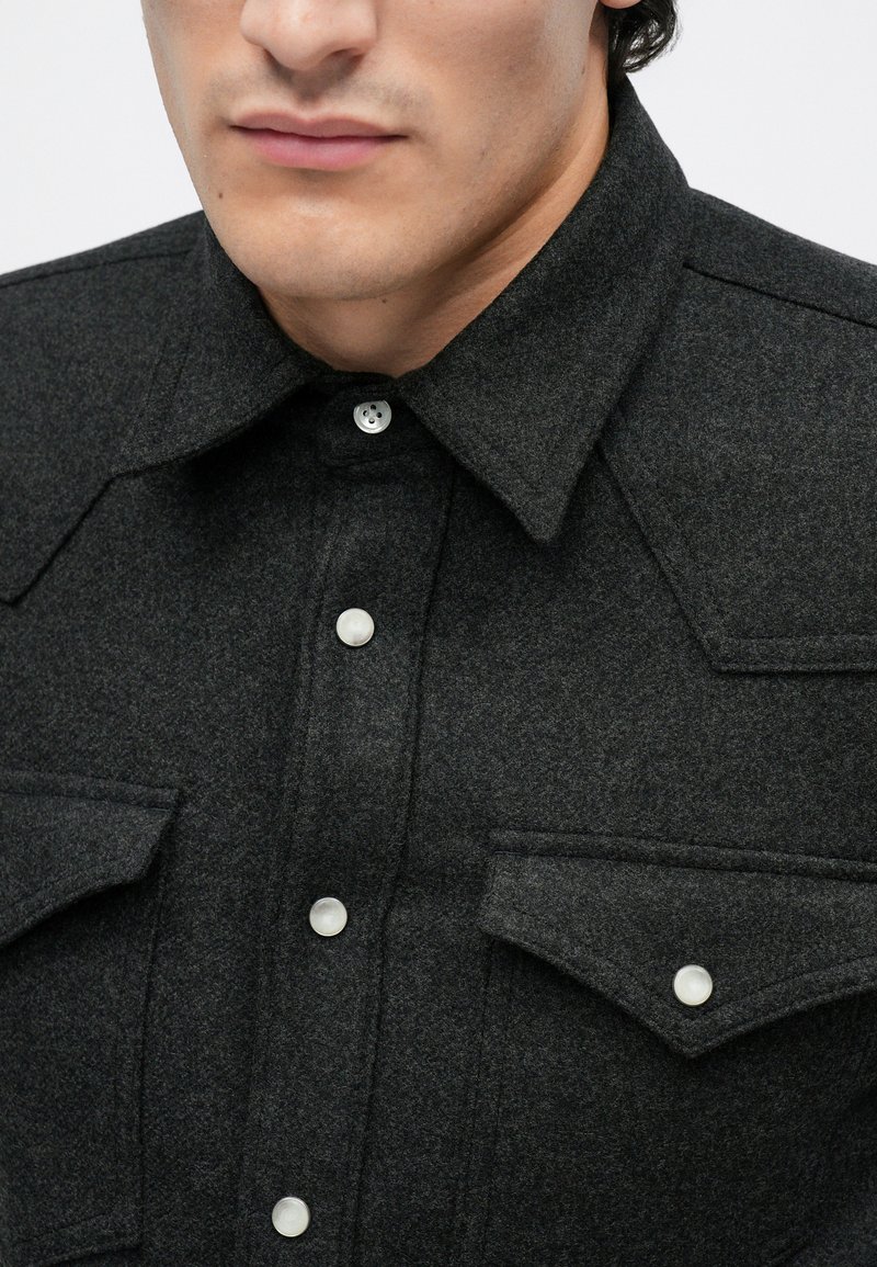 Dark gray wool shirt with a pointed collar, two chest pockets, and white snap buttons for closure. Subtle texture and structured design.