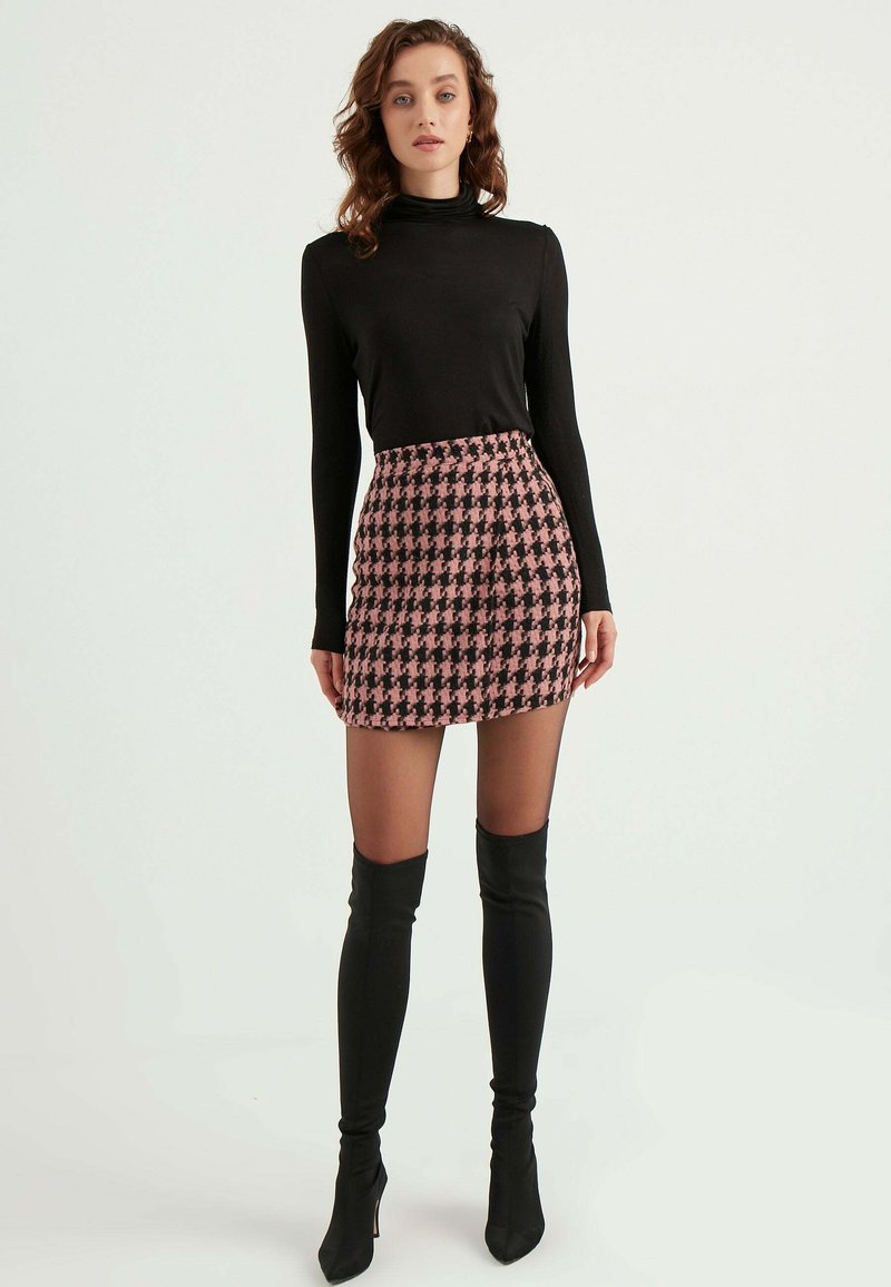 Schwarzes Rollkragenoberteil kombiniert mit einem kurzen Rock im schwarz-pinken Hahnentrittmuster. Das Model trägt knielange schwarze enge Stiefel.