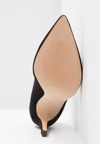 Chaussure à talon haut en daim noir à bout pointu, inclinée vers le haut, montrant une semelle beige clair avec une zone antidérapante texturée.