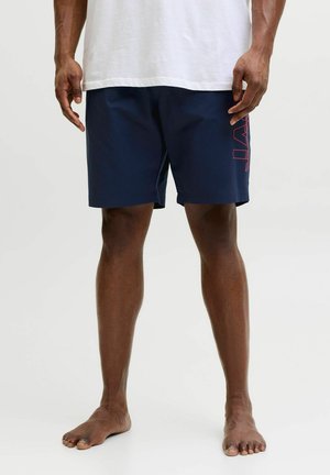 Hombre con pantalones cortos azul marino con texto vertical rojo en el lateral y una camiseta blanca holgada, de pie descalzo sobre un fondo liso.