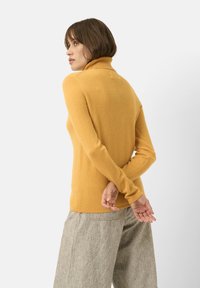 Maglione a collo alto giallo senape in un morbido tessuto testurizzato; design aderente con maniche lunghe, abbinato a pantaloni a gamba larga grigio chiaro.