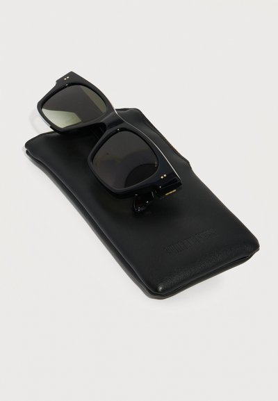 Sorte firkantede solbriller med stel, der ligger på en blød sort læderetui på en hvid baggrund.