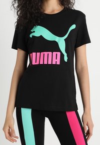 Camiseta negra con logo de Puma en turquesa y letras en rosa. Combinada con mallas negras que tienen franjas turquesa y rosa a lo largo de los lados.