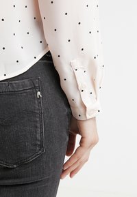 Blouse rose clair à pois noirs, avec manches longues et poignets boutonnés, associée à un jean gris foncé texturé.