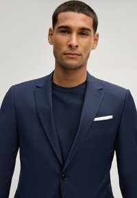 Uomo con capelli corti che indossa un blazer blu navy sopra un maglione blu navy testurizzato, con un fazzoletto bianco nel taschino, su uno sfondo semplice.