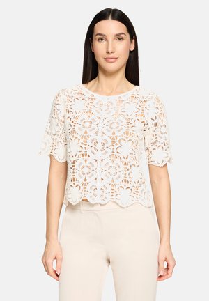 Haut court blanc en dentelle florale avec manches courtes et ourlet festonné, présentant un motif complexe et une texture transparente.