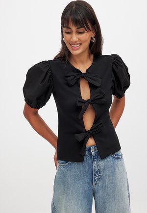 Black blouse featuring puff sleeves, a deep keyhole front, and three fabric bows as closure details. Made of smooth material.