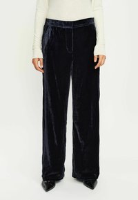 Pantaloni larghi in velluto di un profondo blu navy, caratterizzati da una texture liscia, vita elastica e tasche laterali, abbinati a scarpe nere a punta.