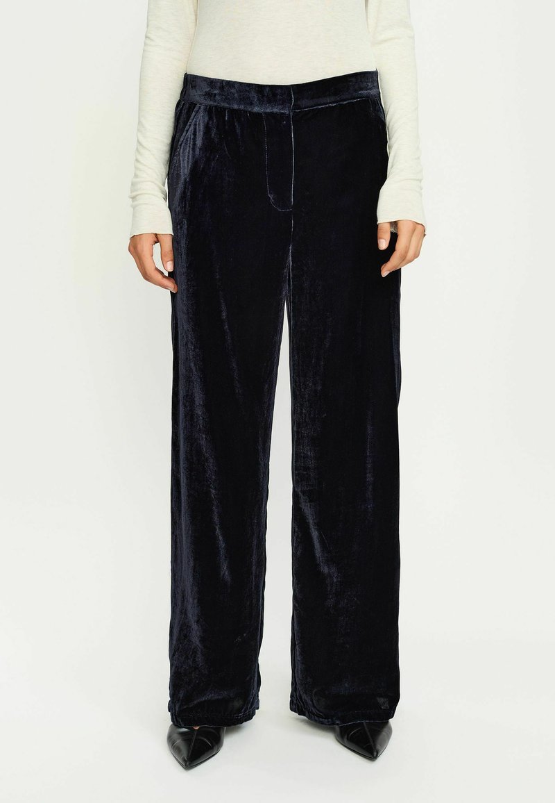 Pantaloni larghi in velluto di un profondo blu navy, caratterizzati da una texture liscia, vita elastica e tasche laterali, abbinati a scarpe nere a punta.