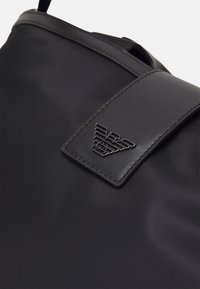 Gros plan d’un sac noir à texture lisse avec un rabat en cuir cousu présentant un petit emblème métallique de logo.