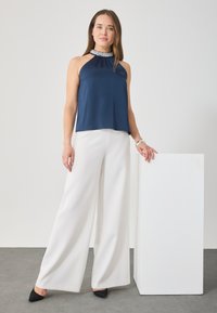 Tengerészkék selyem halter-top gyöngyös gallérral, széles lábú fehér nadrággal és fekete hegyes orrú magassarkúval párosítva, minimalista háttér előtt.