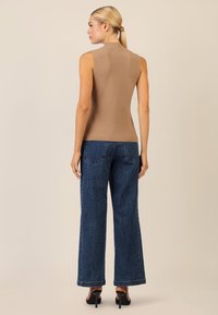 Beige ärmelloses Top mit hohem Kragen, kombiniert mit dunklen, weit geschnittenen Jeans mit dezenten silbernen Nieten und klassischem Fünf-Taschen-Design.