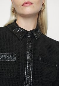 Gros plan d'une personne portant une veste noire texturée avec des boutons noirs brillants, un col et des détails de poche, et des boucles d'oreilles créoles torsadées dorées.