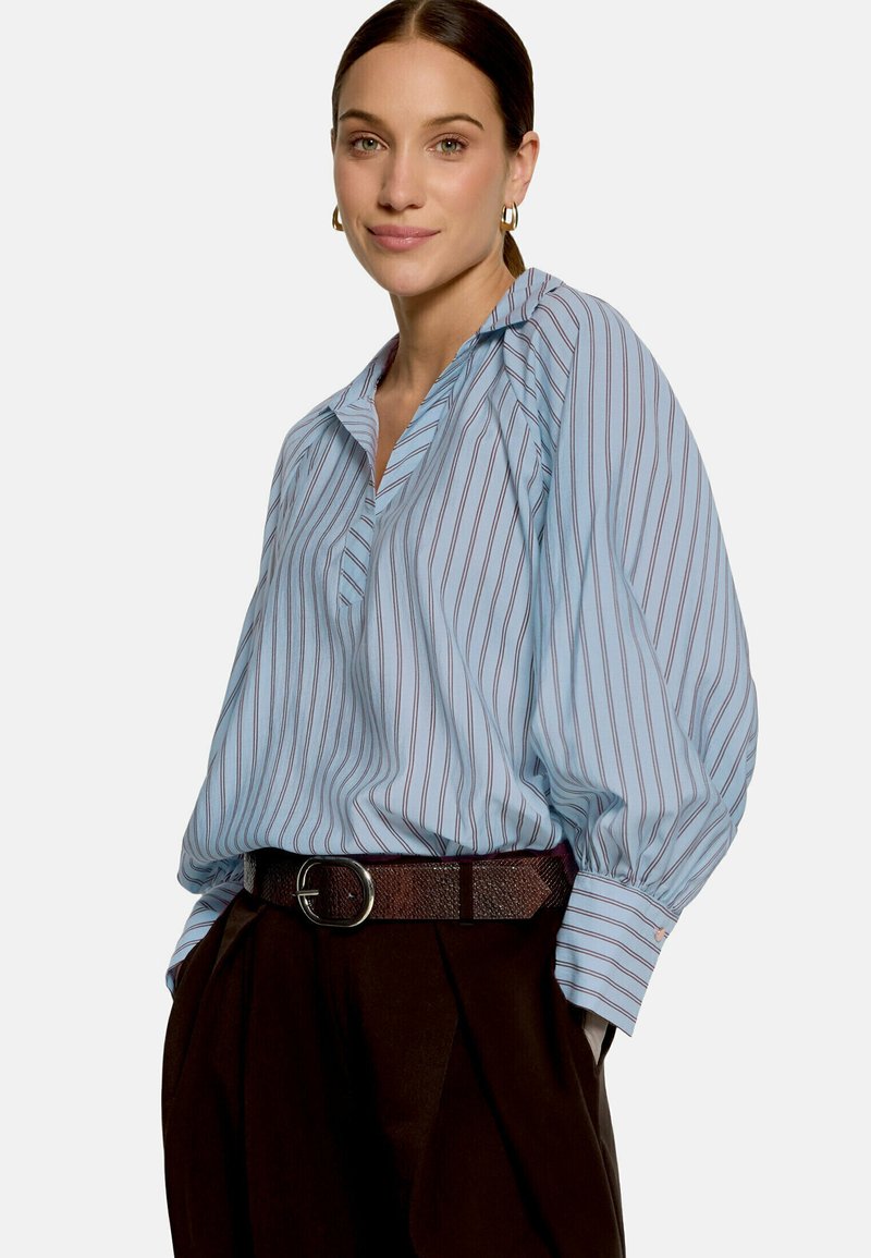 Femme portant un chemisier rayé bleu clair avec des manches bouffantes et un pantalon sombre, souriant légèrement, les cheveux attachés.