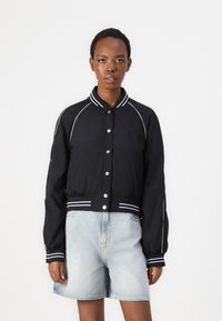 Chaqueta corta negra con ribetes blancos y botones a presión. Presenta mangas largas y un cuello acanalado. Se lleva con pantalones cortos de mezclilla azul claro.