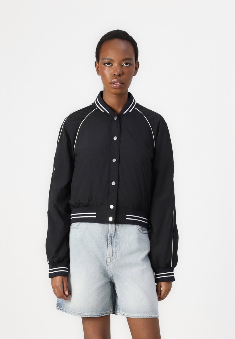 Chaqueta corta negra con ribetes blancos y botones a presión. Presenta mangas largas y un cuello acanalado. Se lleva con pantalones cortos de mezclilla azul claro.