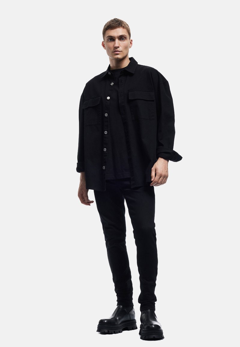 Camisa negra de botones con dos bolsillos en el pecho, llevada sobre una camiseta negra y combinada con jeans skinny negros y botines negros gruesos.