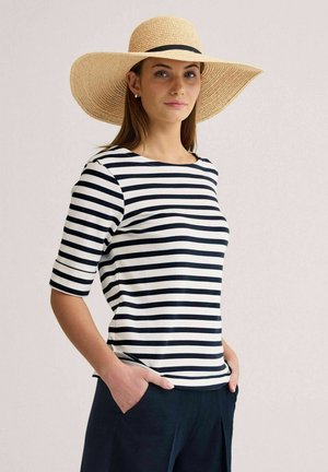 Nainen, jolla on päällään mustavalkoraidallinen paita, tummansiniset housut ja leveälierinen olkihattu, seisoo käsineet taskuissa vaaleaa taustaa vasten.