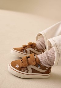 Barna velúr sportcipő fehér részletekkel, két tépőzáras pánttal és hímzett kutya mintákkal. Bordázott, világos színű zoknival viselve.