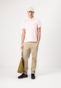 Camisa polo rosa, chinos beige, zapatillas negras y una gorra color crema. Sostiene una bolsa de mano verde oliva. Líneas limpias, ajuste casual, marca mínima.