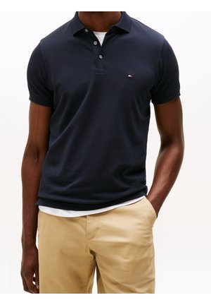 Mann trägt ein dunkelblaues Poloshirt über einem weißen Unterhemd und beige Hose, eine Hand in der Tasche, vor hellem einfarbigem Hintergrund.