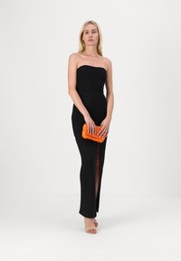 Črna brezramna obleka z bočnim razporkom, izdelana iz teksturirane tkanine. Manekenka drži živahno oranžno torbico s sequins in nosi golenke s peto.