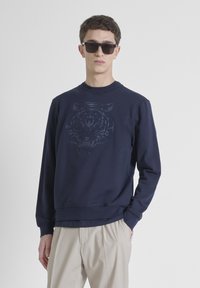 Sudadera azul marino con un gráfico de tigre texturizado en la parte frontal, cuello redondo, puños y dobladillo de canalé. Se lleva con pantalones beige.