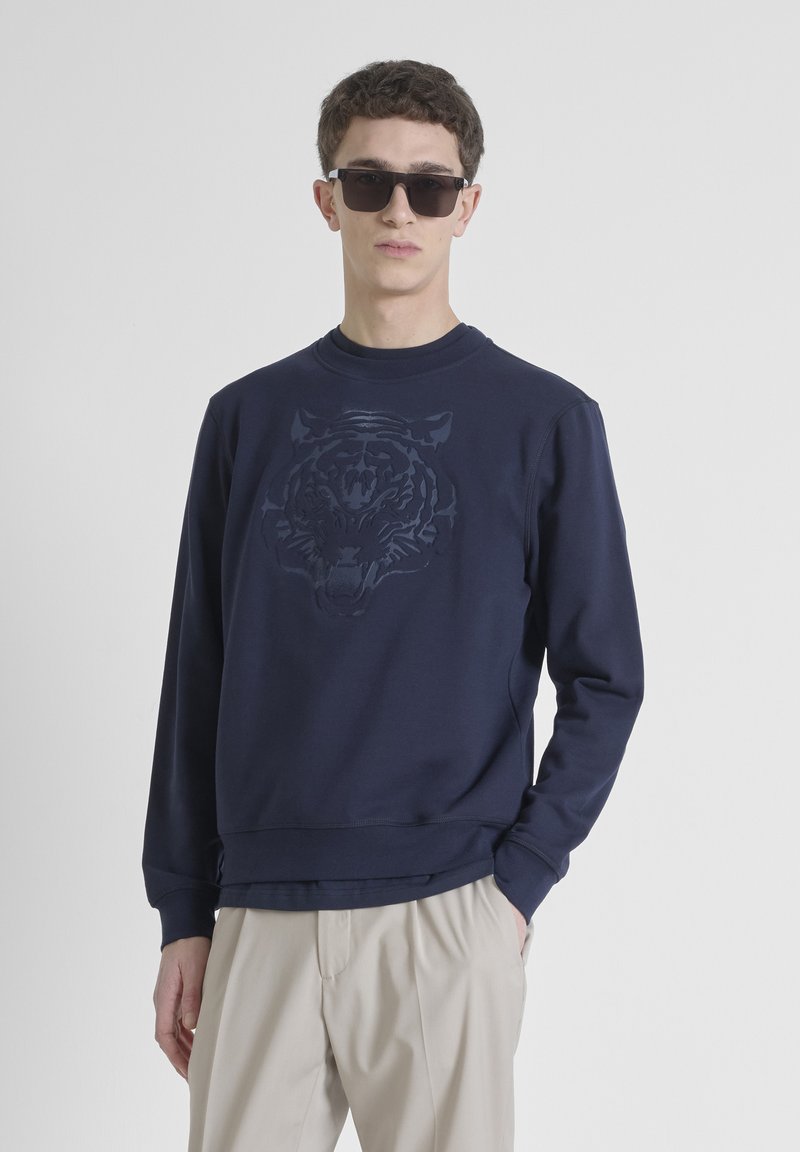 Felpa blu navy con una grafica di un tigre testurizzata sul davanti, colletto a girocollo, polsini e orlo a coste. Indossata con pantaloni beige.