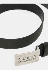 Schwarzer Gummigürtel mit glatter Textur und einer silberfarbenen Metallschnalle, die mit dem eingeprägten Text "GUESS LOS ANGELES" versehen ist. Verstellbar mit Löchern.