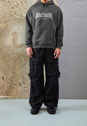 Persona con una sudadera con capucha gris oscuro con el texto "Vertex Berlin" y pantalones cargo negros holgados con múltiples bolsillos, de pie frente a una pared texturizada.