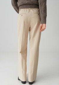 Beige op maat gemaakte broek met een ontspannen pasvorm, met een vlakke voorkant, achterzakken met knoopdetails en een soepele, lichtgewicht stof.
