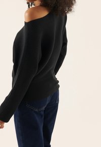 Femme aux cheveux bouclés portant un pull noir côtelé à épaules dénudées et un jean bleu foncé, sur un fond clair uni.