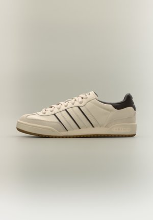 Кремава ниска маратонка Adidas с три тъмни ивици отстрани, черен панел на петата, бели връзки и подметка в цвят на гума, показана в профил.