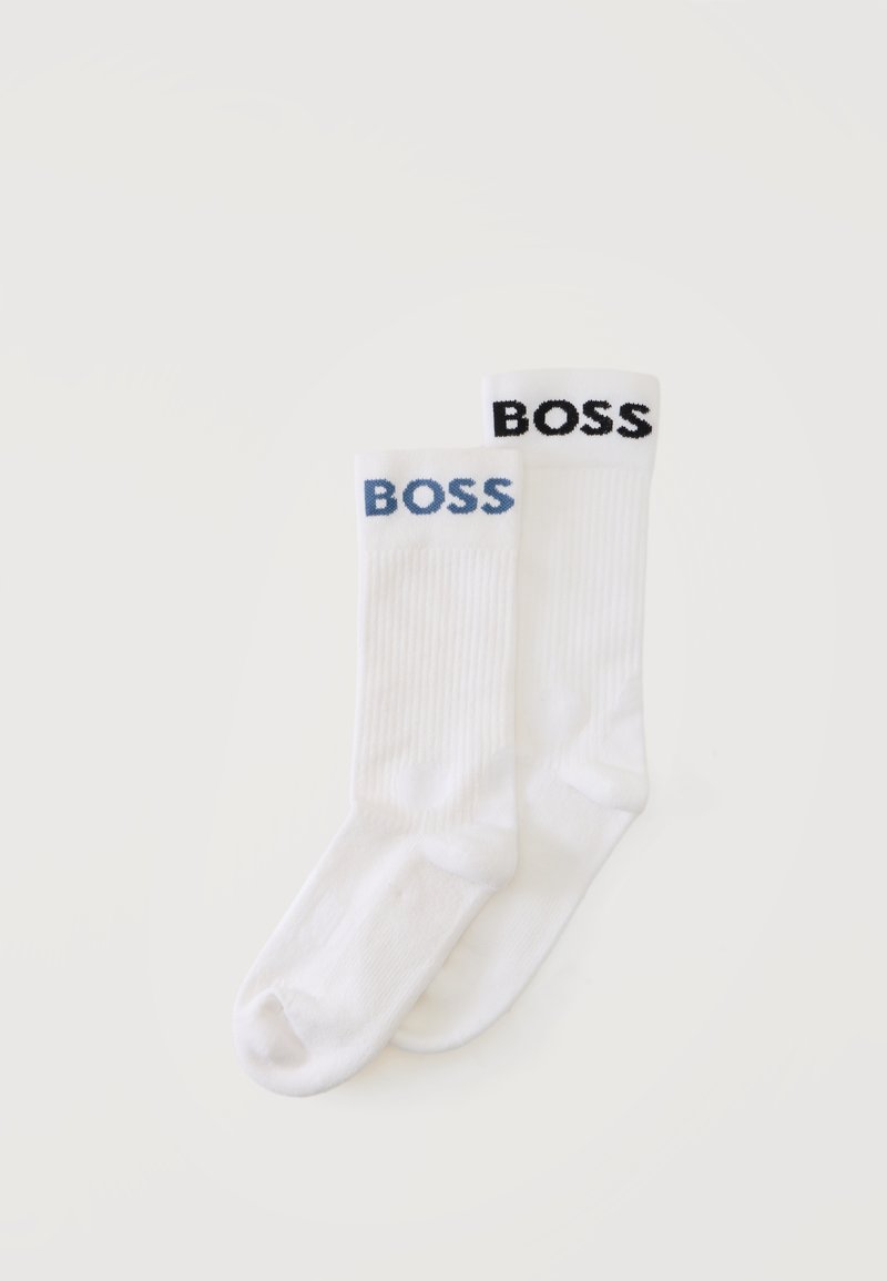 Deux chaussettes montantes blanches avec une texture côtelée, une avec le texte "BOSS" en bleu et l'autre en noir sur le haut du bord, sur un fond clair uni.