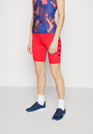 Κόκκινες ποδηλατικές σορτσάκια με μαύρες λεπτομέρειες του λογότυπου Kappa, συνδυασμένα με ένα πολυχρωμο tank top με σχέδια και ναυτικά αθλητικά παπούτσια. Ορατές λευκές κάλτσες αστραγάλου.