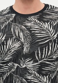 Άνδρας που φοράει μαύρο μπλουζάκι με λαιμόκοψη crew με λευκό και γκρι μοτίβο φύλλων φοίνικα, εμφανίζονται μόνο ο λαιμός και το πάνω μέρος του στήθους.