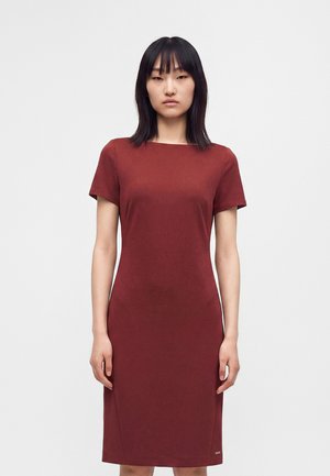 Femme aux cheveux noirs lisses portant une robe ajustée, à manches courtes, longueur genou, rouge foncé, se tenant devant un fond clair uni.