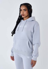 Lichtgrijze hoodie met een voorzak, een trekkoord en een wit logo op de borst, gecombineerd met bijpassende aansluitende joggers. Zachte stoffen textuur.