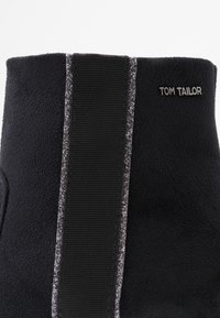 Close-up van de bovenkant van een zwarte stoflaars met een verticale elastische zwarte strook en het "TOM TAILOR" logo aan de zijkant.
