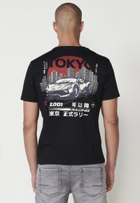 Melna īsām piedurknēm T-kreklā uz muguras ir grafikā attēlots sacīkšu auto un Tokijas debesskrāpjus sarkanā un baltā krāsā. Izgatavots no kokvilnas materiāla.