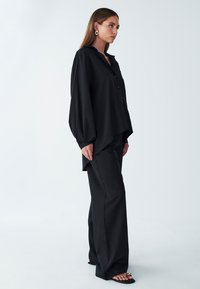 Camicia nera oversize con bottone frontale, maniche lunghe larghe e orlo asimmetrico, abbinata a pantaloni larghi coordinati e sandali piatti.