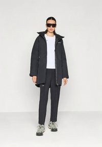 Svart vanntett jakke med hette, frontglidelås og sidelommer, brukt over en hvit skjorte og mørke bukser. Modell bærer også mørke solbriller.