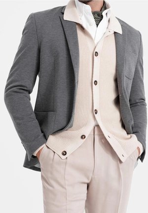 Hombre vistiendo un cárdigan de punto beige en capas, camisa blanca, bufanda estampada, blazer gris texturizado y pantalones beige, con las manos en los bolsillos.