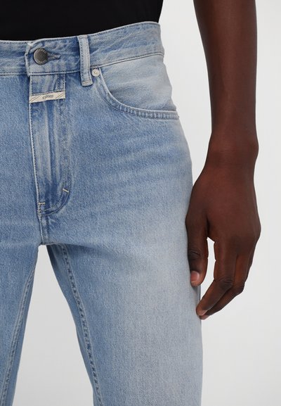 Jeans en denim bleu clair à coupe slim, présentant un design classique à cinq poches, une fermeture par bouton et zip, et des coutures contrastées.