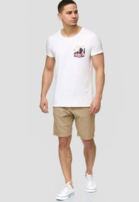 White cotton t-shirt with a pink palm tree print pocket, paired with beige shorts and white sneakers. Model stands against a neutral background.