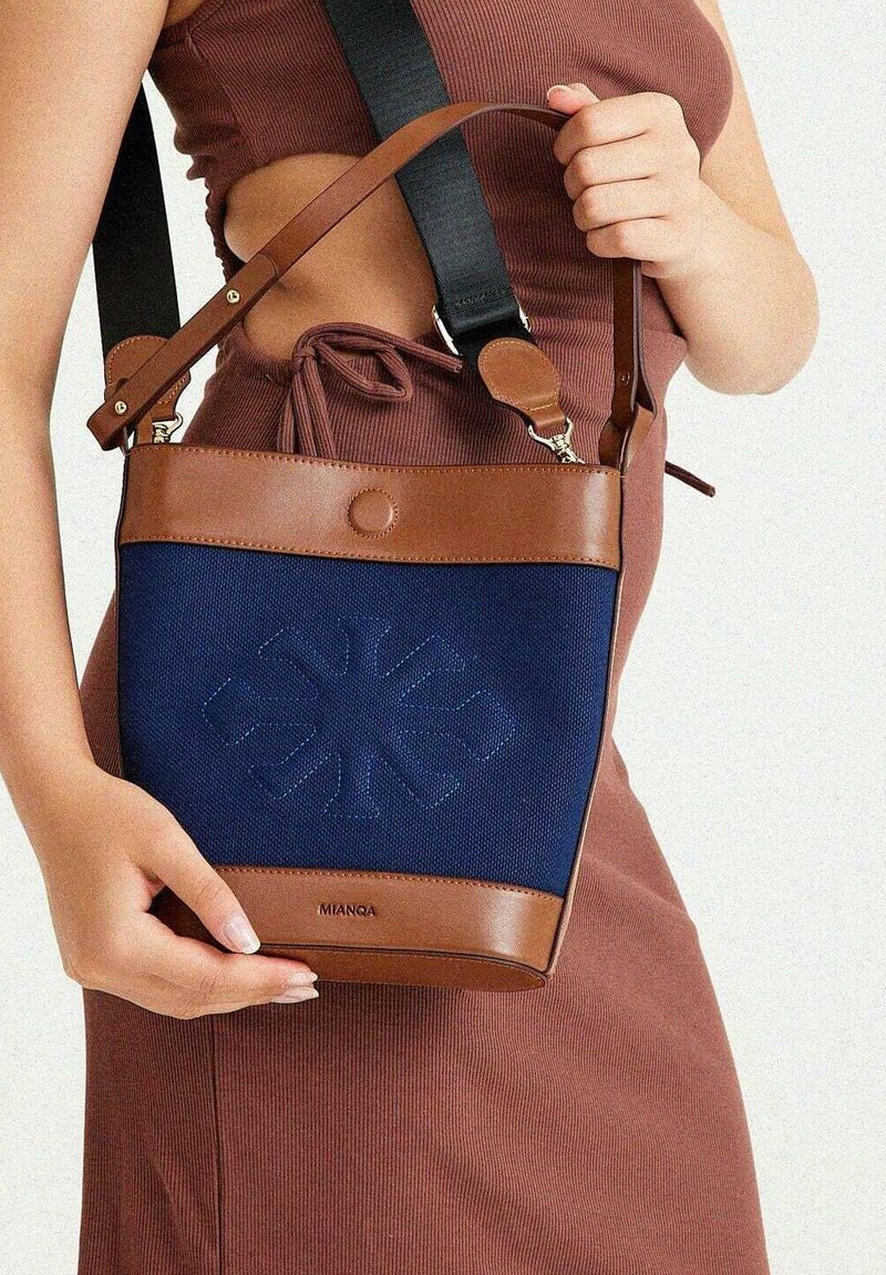 Navy blue canvas bag with brown leather accents, adjustable strap, and embossed floral pattern. Compact size with a top snap closure.