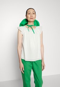 Blusa branca de manga curta com decote franzido e detalhes em verde, combinada com calças verdes. A blusa apresenta um acabamento de renda e mangas com babados.