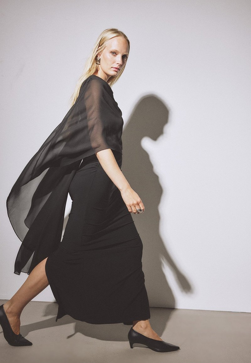 Blonde woman walking in a long black dress with sheer overlay and low black heels against a white background casting a shadow.